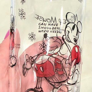 Pu&ograve; includere: Bicchiere di vetro trasparente con un'illustrazione di Topolino in nero, rosso e bianco. Il design include fiocchi di neve e il testo "Mickey can have shoulders when needed". Un bicchiere decorativo.