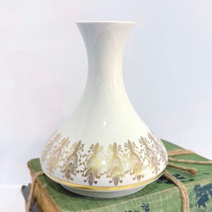 Può includere: Un vaso di ceramica bianca con un motivo floreale dorato attorno alla base. Il vaso è appoggiato su una pila di libri vintage con una copertina verde e un legaccio di spago.