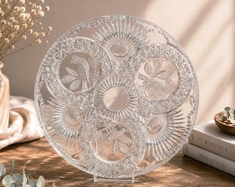 May include: A clear, round glass serving platter with intricate etched designs. The platter features detailed patterns, including birds and leaves, and is supported by a clear stand. The design is symmetrical and elegant.