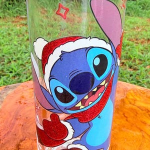 May include: Clear glass tumbler featuring a blue cartoon character wearing a Santa hat and red scarf. The character has large black eyes and a wide smile. The tumbler is decorated with red and white accents, perfect for holiday drinks.