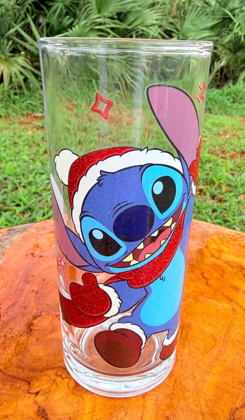May include: Clear glass tumbler featuring a blue cartoon character wearing a Santa hat and red scarf. The character has large black eyes and a wide smile. The tumbler is decorated with red and white accents, perfect for holiday drinks.