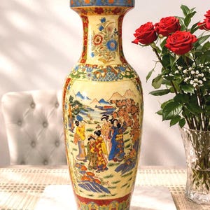May include: A tall, decorative ceramic vase with a cream-colored background and a detailed Asian-inspired design. The vase features a painted scene of figures in traditional clothing, with red, blue, and gold accents. The vase is approximately 18 inches tall.