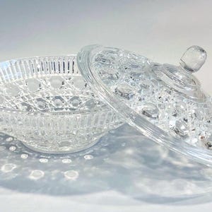 May include: A clear, cut-glass candy dish with a lid. The dish has a round shape with a decorative pattern and a small knob on the lid. The lid is partially open, revealing the interior of the dish. The dish is sitting on a white surface.