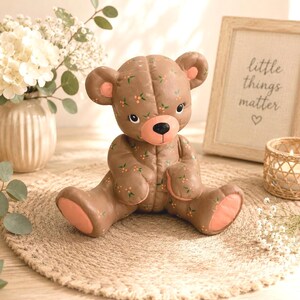 Può includere: Una statuina di orsetto marrone con un motivo floreale e dettagli pesca. L'orso è seduto su un tappetino rotondo di iuta. Un cartello incorniciato con la scritta "little things matter" è sullo sfondo.