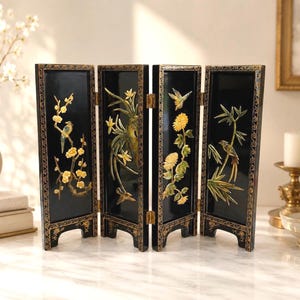 May include: A four-panel folding screen featuring a glossy black finish with gold-painted border patterns. Each panel displays raised, multi-colored relief carvings of birds, flowers, and bamboo stalks, connected by small gold-toned metal hinges.