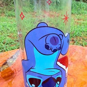 May include: A clear glass tumbler featuring a blue cartoon character, with a large head and big eyes. The character is upside down, with a red and white design at the bottom. Red star and square designs are also on the glass.
