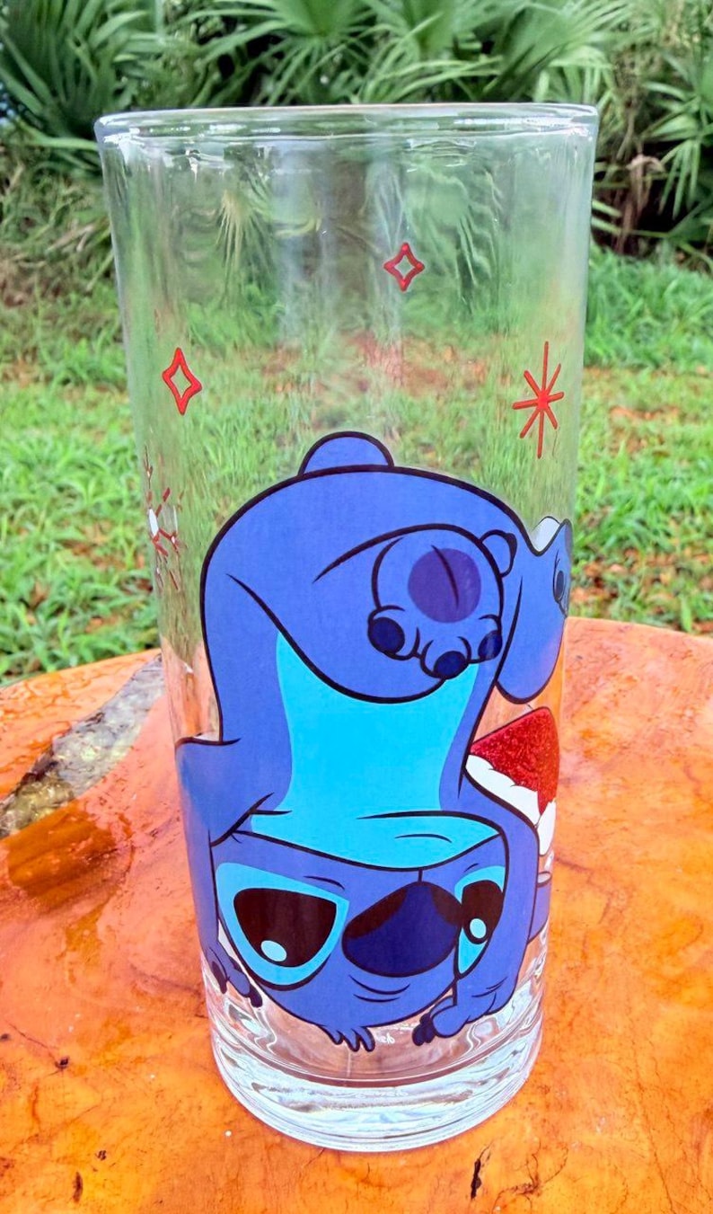 May include: A clear glass tumbler featuring a blue cartoon character, with a large head and big eyes. The character is upside down, with a red and white design at the bottom. Red star and square designs are also on the glass.