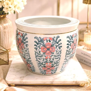 May include: A white ceramic planter with a rounded silhouette, featuring a repeating vertical pattern of pink flowers and green leafy vines. The rim is accented with a thin gold line, and the surface is decorated with delicate, hand-painted floral motifs.