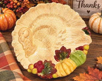 May include: A round ceramic plate featuring a raised, embossed turkey design in a cream colour. The bottom edge is decorated with colourful, three-dimensional painted accents including a yellow pumpkin, a green pumpkin, red grapes, and dark red leaves.