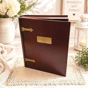 May include: A dark brown wooden photo album with brass hardware and a brass nameplate. The album is open, revealing the inside pages. The background includes a vase of white flowers, a framed sign that says "Collect Moments Not Things", and a white lantern.