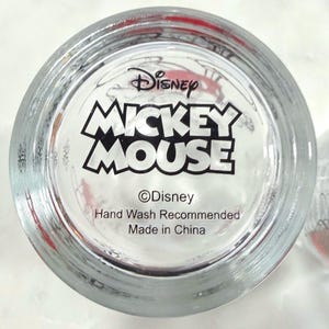 Pu&ograve; includere: Bicchiere trasparente con le parole "Disney MICKEY MOUSE" in bianco e nero. Il fondo del bicchiere include anche il logo Disney e il testo "Hand Wash Recommended Made in China."