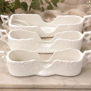 May include: Three white ceramic sleigh-shaped dishes arranged in a stack. Each dish features an elongated, open-top design with ornate, embossed floral and scrollwork detailing along the exterior rims and two small, decorative handles on each end.