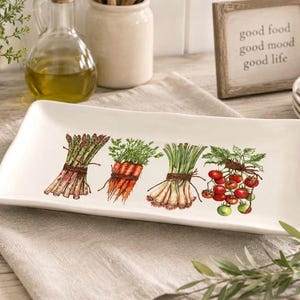Puede incluir: Una bandeja de servir rectangular de cerámica blanca con una ilustración colorida de cuatro manojos de verduras: espárragos, zanahorias, cebolletas y tomates rojos, cada uno atado con un cordel marrón sobre la superficie lisa.