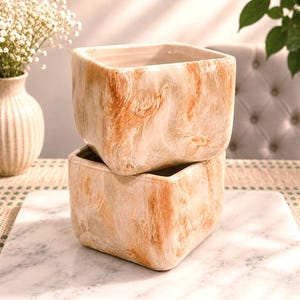 May include: Two square ceramic planters stacked on a marble surface. The planters have a marbled beige and orange pattern. A vase with white flowers is visible in the background.