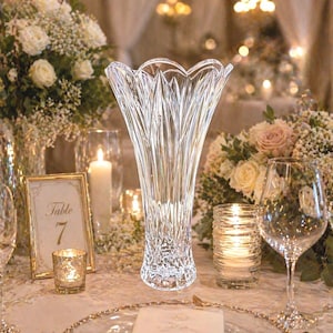 May include: A clear, cut-glass vase with a scalloped rim stands on a table set for a special occasion. The vase has a textured, vertical pattern. The table setting includes flowers, candles, and glassware.