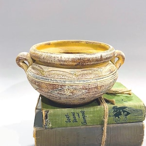 Può includere: Una ciotola decorativa in ceramica invecchiata con due manici, con una finitura bianca e marrone anticata. La ciotola è appoggiata su due libri verdi vintage legati con spago. La ciotola ha un interno giallo e intagli dettagliati.