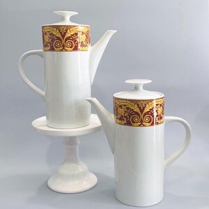 May include: Two white ceramic coffee pots with ornate gold and burgundy detailing on the lids. One pot sits on a small white pedestal. The pots have a classic, elegant design, suitable for serving coffee or tea.