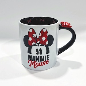 Puede incluir: Taza de cer&aacute;mica blanca con Minnie Mouse. La taza tiene un asa e interior negros, con un lazo rojo en el asa. La parte delantera muestra la cara de Minnie, un lazo rojo con lunares blancos y las palabras "MINNIE Mouse" en negro y rojo.