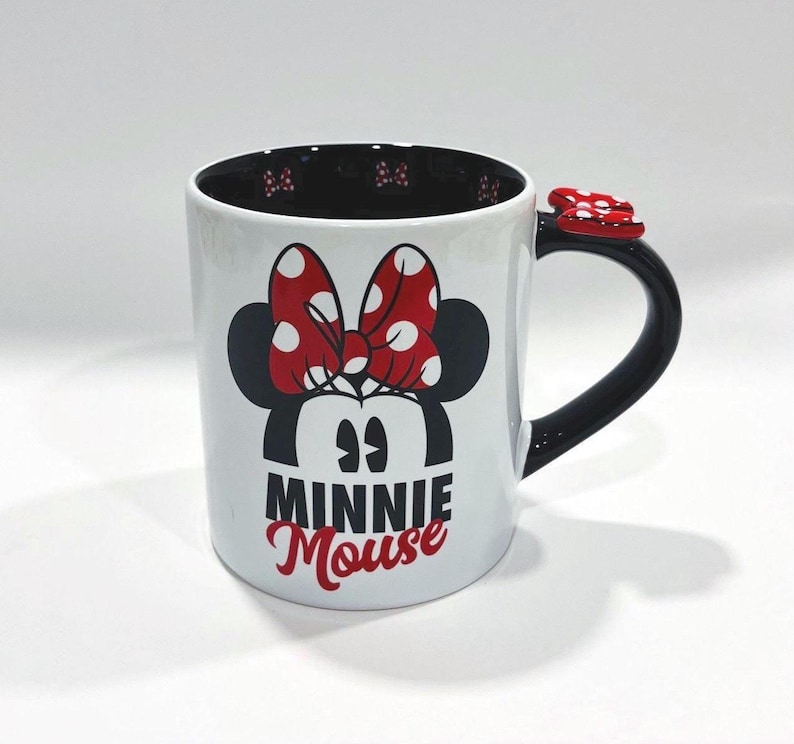 Puede incluir: Taza de cer&aacute;mica blanca con Minnie Mouse. La taza tiene un asa e interior negros, con un lazo rojo en el asa. La parte delantera muestra la cara de Minnie, un lazo rojo con lunares blancos y las palabras "MINNIE Mouse" en negro y rojo.