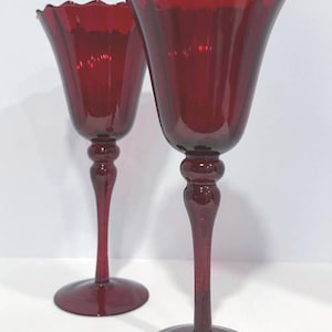 May include: Two ruby red wine glasses with a scalloped rim. The glasses have a bulbous stem and a round base. The glasses are made of glass and are set against a white background.