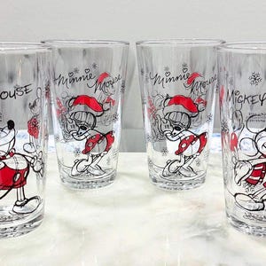 Pu&ograve; includere: Quattro bicchieri trasparenti con disegni festivi di Topolino e Minnie. Ogni bicchiere mostra un'illustrazione diversa dei personaggi in abiti rossi e bianchi, con le parole "Mickey Mouse" e "Minnie Mouse" sul bicchiere.