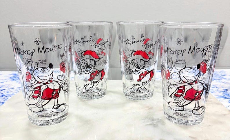 Pu&ograve; includere: Quattro bicchieri trasparenti con disegni festivi di Topolino e Minnie. Ogni bicchiere mostra un'illustrazione diversa dei personaggi in abiti rossi e bianchi, con le parole "Mickey Mouse" e "Minnie Mouse" sul bicchiere.