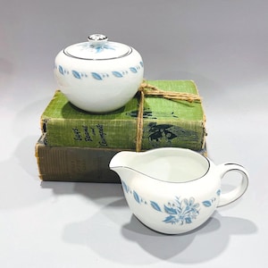 May include: A white porcelain sugar bowl with a lid and a matching creamer. Both pieces feature a delicate blue floral pattern and black trim. The sugar bowl sits atop a stack of vintage green books.