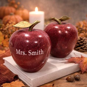 May include: Two decorative red apple sculptures with gold-colored leaves and stems. One apple has the name "Mrs. Smith" engraved on it. The apples are on a book, with a lit candle and fall foliage in the background.