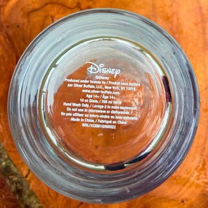 May include: Clear glass with the Disney logo and product information printed on the bottom. The text includes the manufacturer's details, capacity of 10 oz, and care instructions. The glass is made in China.