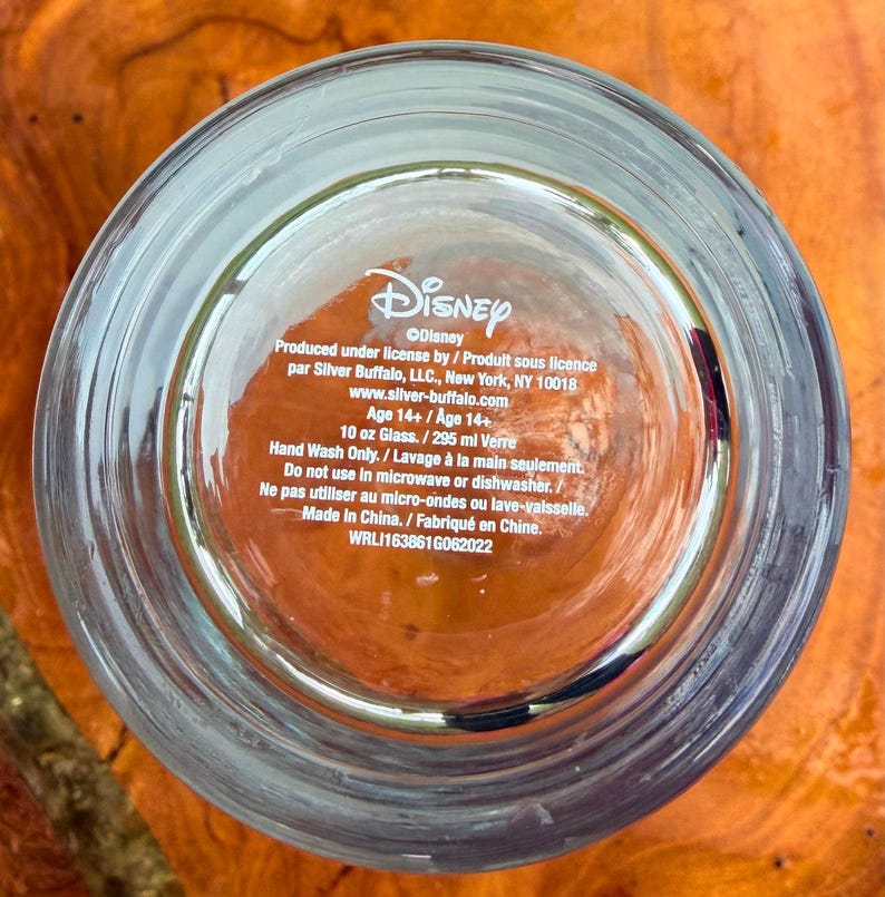 May include: Clear glass with the Disney logo and product information printed on the bottom. The text includes the manufacturer's details, capacity of 10 oz, and care instructions. The glass is made in China.