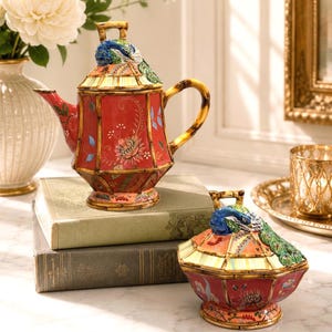 May include: A ceramic teapot and matching lidded sugar bowl featuring a red base with floral patterns and bamboo-style framing. Each lid is topped with a sculpted, colorful peacock figure with detailed plumage and a handle shaped like bamboo.