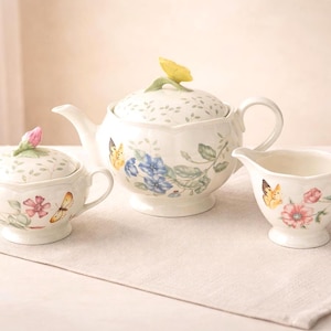 May include: A white porcelain tea set featuring a teapot, sugar bowl, and creamer. The set is decorated with floral and butterfly motifs in shades of pink, blue, and yellow. The teapot has a yellow flower on the lid.