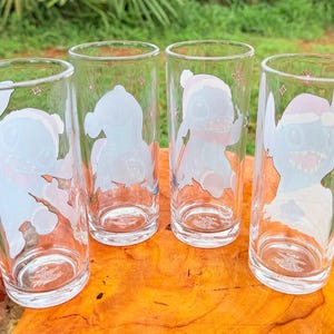 May include: Four clear drinking glasses with cartoon character designs. Each glass features a different image of a blue alien-like creature with white and pink accents. The glasses are cylindrical and sit on a wooden surface.