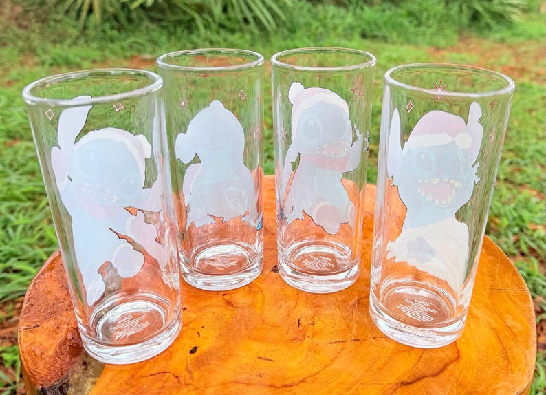 May include: Four clear drinking glasses with cartoon character designs. Each glass features a different image of a blue alien-like creature with white and pink accents. The glasses are cylindrical and sit on a wooden surface.
