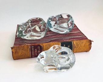 Puede incluir: Tres candelabros transparentes de cristal con forma de conejito sobre un libro antiguo con tapa granate. Los candelabros son transparentes. El lomo del libro es visible, con el título "HISTORY OF THE WORLD".