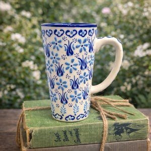 May include: Tall ceramic mug with a white handle and a blue floral pattern. The mug is decorated with blue flowers and leaves on a white background. The mug is sitting on top of two green books tied together with twine.