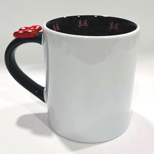 Puede incluir: Taza de cer&aacute;mica blanca con asa negra y un lazo rojo. El interior de la taza es negro con patrones de lazos rojos. El adorno de lazo es rojo con lunares blancos.