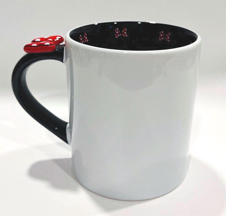 Puede incluir: Taza de cer&aacute;mica blanca con asa negra y un lazo rojo. El interior de la taza es negro con patrones de lazos rojos. El adorno de lazo es rojo con lunares blancos.