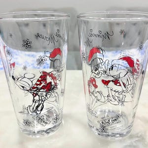 Pu&ograve; includere: Due bicchieri trasparenti con illustrazioni di Minnie e Topolino con cappelli da Babbo Natale. I bicchieri sono decorati con fiocchi di neve e la scritta "Minnie Mouse" in corsivo.