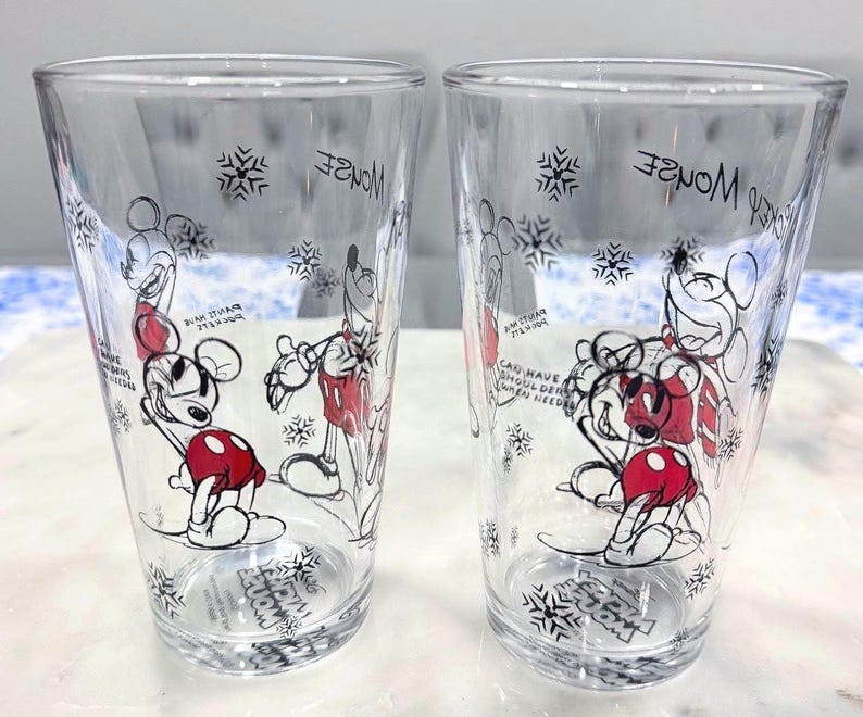 Pu&ograve; includere: Due bicchieri trasparenti con un design di Topolino. I bicchieri presentano un'illustrazione di Topolino in bianco e nero e rosso, fiocchi di neve e testo. Il testo include "Mickey Mouse" e "Can have shoulder when needed."