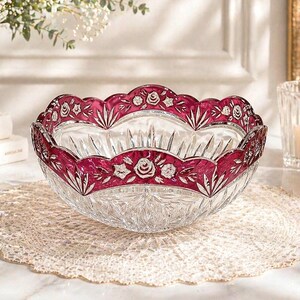 May include: A clear crystal bowl with a scalloped, ruby red rim. The rim features white floral and leaf designs. The bowl sits on a cream-colored, lace doily. A candle in a glass holder is in the background.