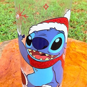 May include: A clear glass featuring a cartoon character, Stitch, wearing a Santa hat and scarf. The character is blue with large eyes and a wide smile. A red snowflake and a small red square are also on the glass.
