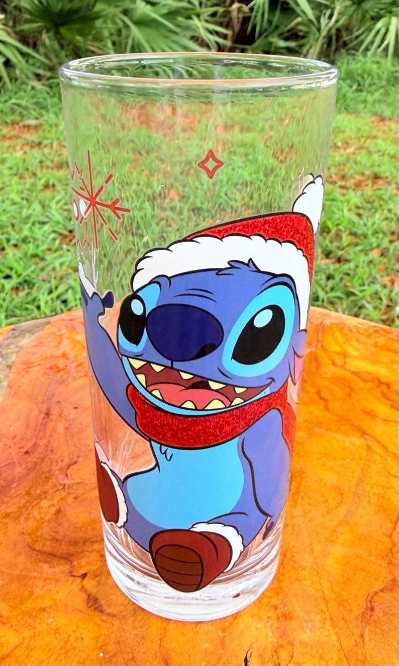 May include: A clear glass featuring a cartoon character, Stitch, wearing a Santa hat and scarf. The character is blue with large eyes and a wide smile. A red snowflake and a small red square are also on the glass.