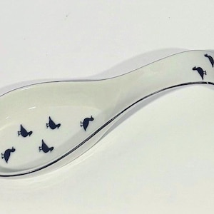 May include: A white ceramic spoon rest with a curved design and a black trim. The spoon rest is decorated with a pattern of small, black bird silhouettes. This kitchenware item is designed to hold spoons and other cooking utensils.