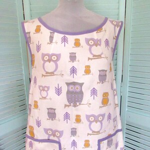 May include: A white apron with a purple trim and a pattern of owls and trees. The apron has two pockets.