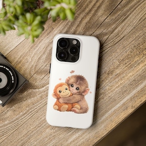 May include: White phone case featuring a cute illustration of two hugging monkeys. One monkey is light brown, the other is a darker brown. Small pink hearts are scattered around the monkeys. The phone case has a black camera module.