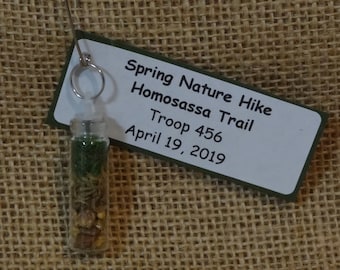 May include: A small glass bottle filled with dried plants and seeds, attached to a safety pin. A white label with green trim reads "Spring Nature Hike Homosassa Trail Troop 456 April 19, 2019".