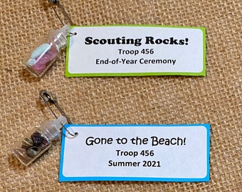 May include: Two small glass bottles with cork tops and safety pins attached. The bottles contain small rocks and shells. The bottles are attached to cards with text that reads "Scouting Rocks! Troop 456 End-of-Year Ceremony" and "Gone to the Beach! Troop 456 Summer 2021".