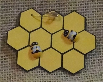 May include: A yellow honeycomb-shaped pin with two black and yellow bees on it. The pin is attached to a safety pin.