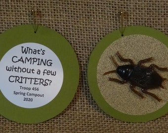 May include: Two green and white round paper ornaments. One ornament has the text "What's CAMPING without a few CRITTERS? Troop 456 Spring Campout 2020". The other ornament has a brown plastic cockroach on a tan background.
